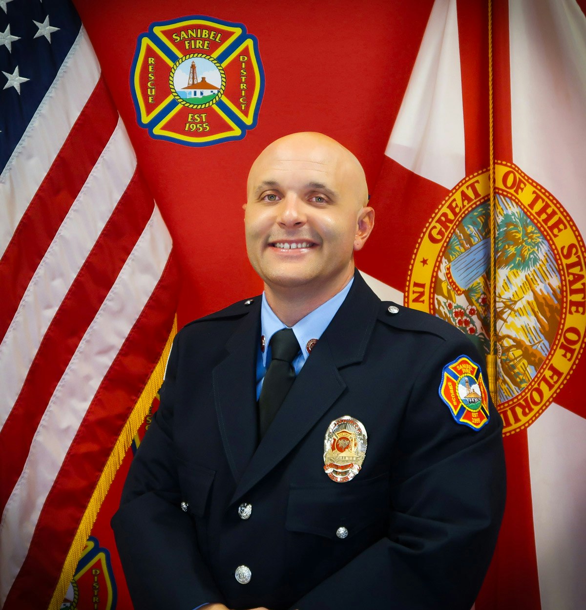 A firefighter in uniform stands smiling in front of American and Florida flags, and a Sanibel Fire Rescue District emblem.