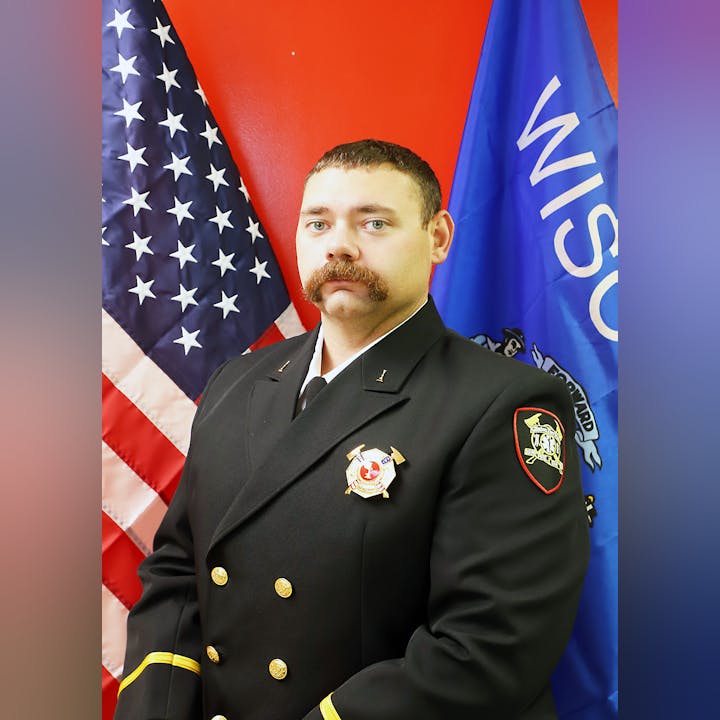 A person in a formal uniform with badges, standing before the U.S. and Wisconsin flags.