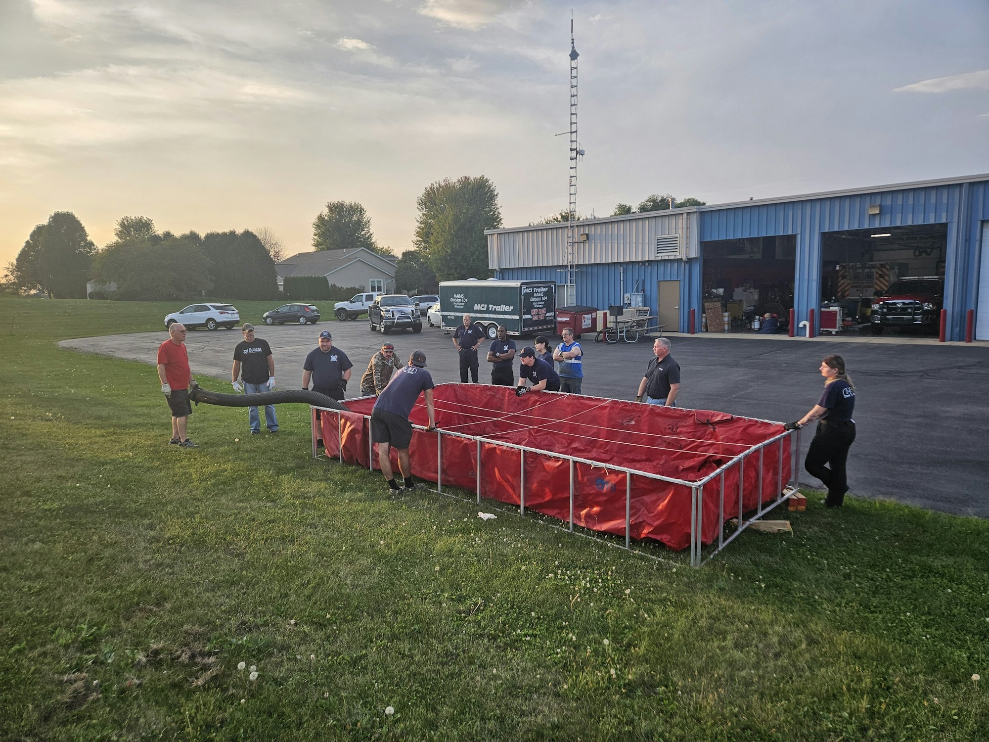 A group of people sets up a large, red portable tank by a building, possibly for firefighting or training.