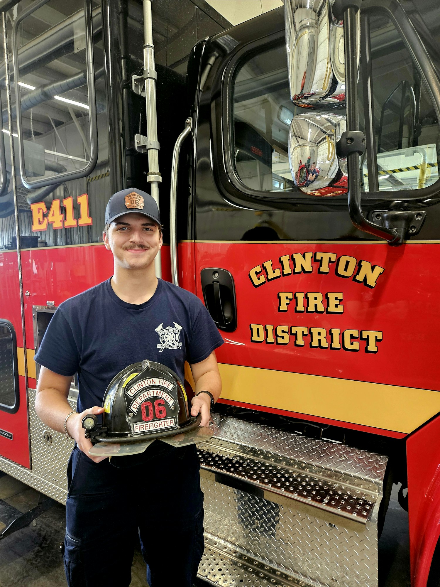 A person holding a firefighter helmet stands in front of a red and black fire truck labeled "Clinton Fire District."