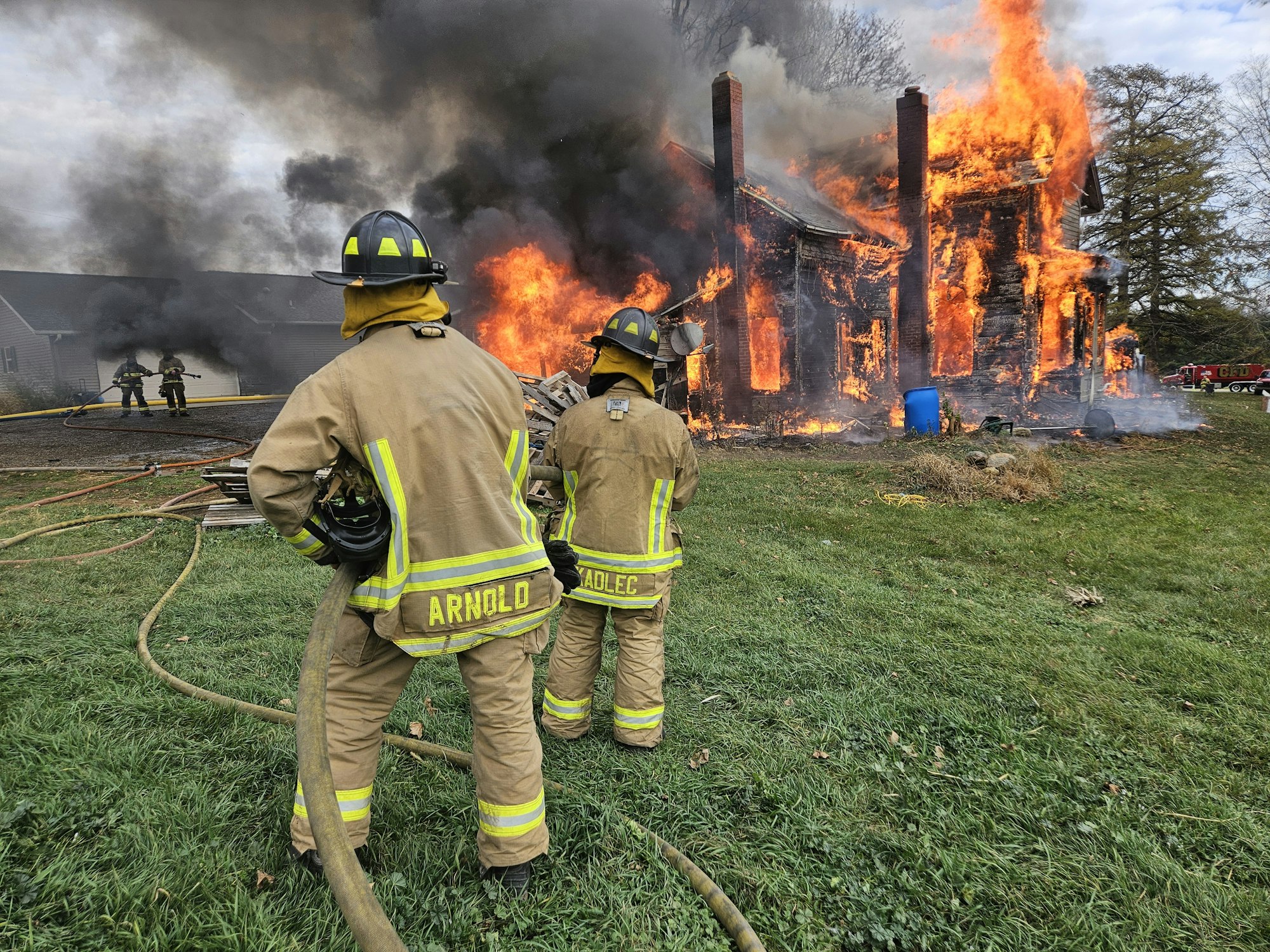 Firefighters in gear watching a house engulfed in flames.