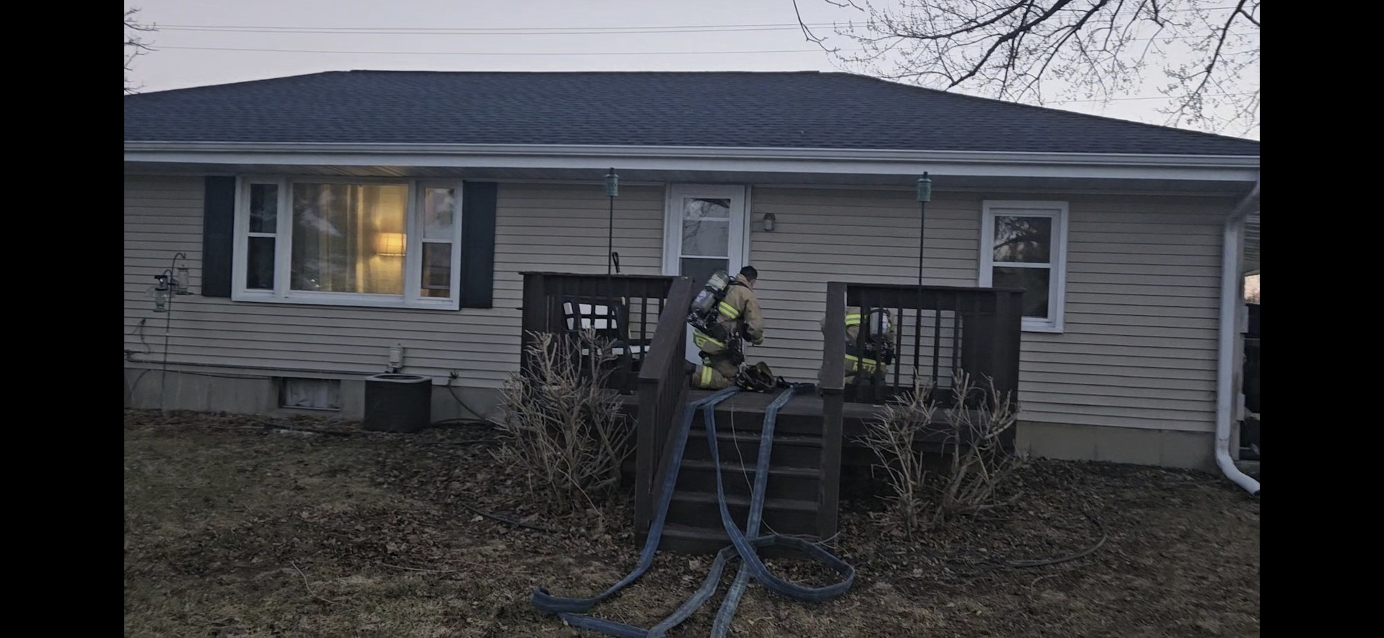 Firefighters in gear are at a house, with hoses on the ground, responding to an incident outside.