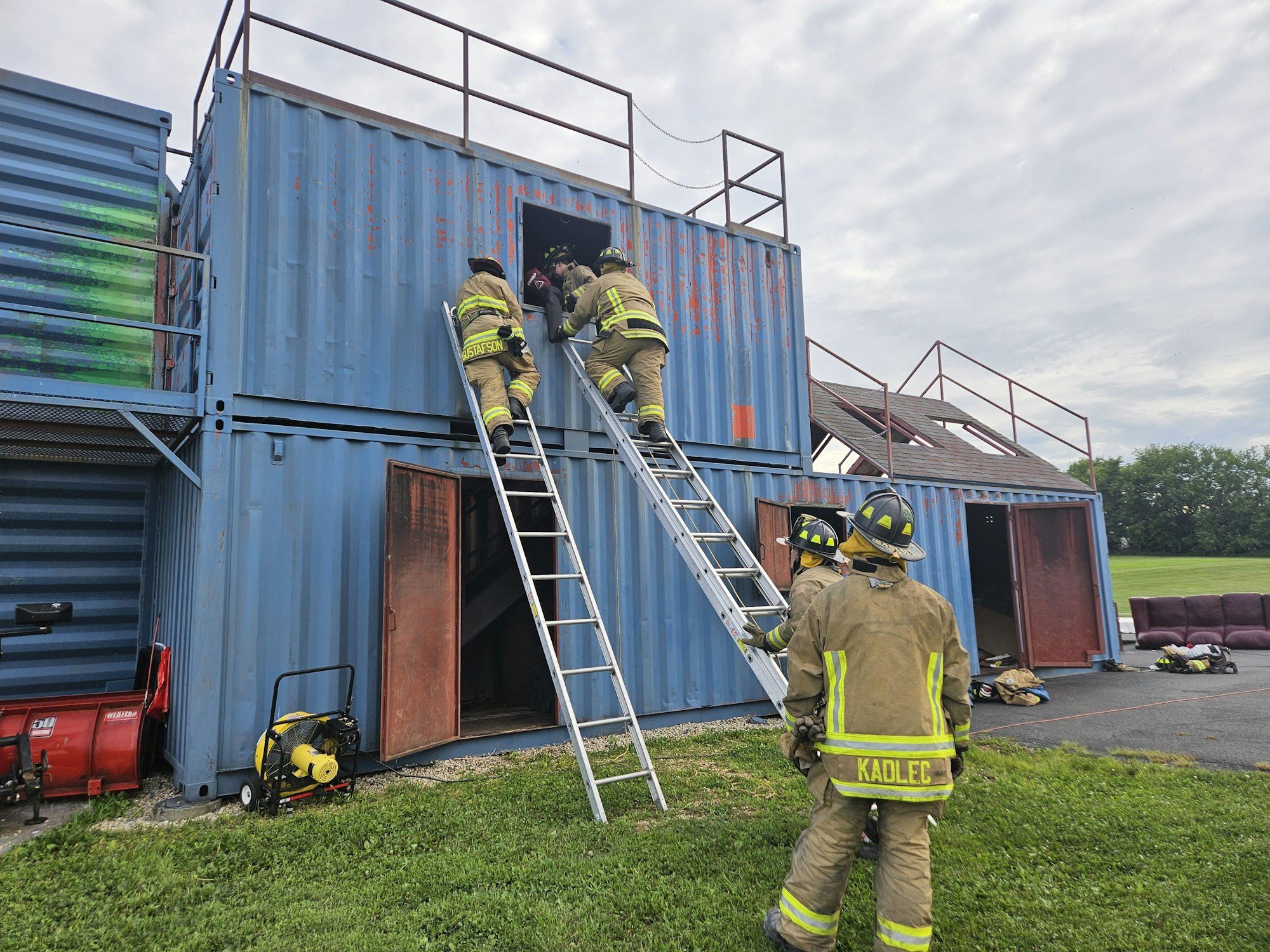 Firefighters in gear climb ladders into a two-story training structure made of shipping containers, with firefighting equipment nearby.