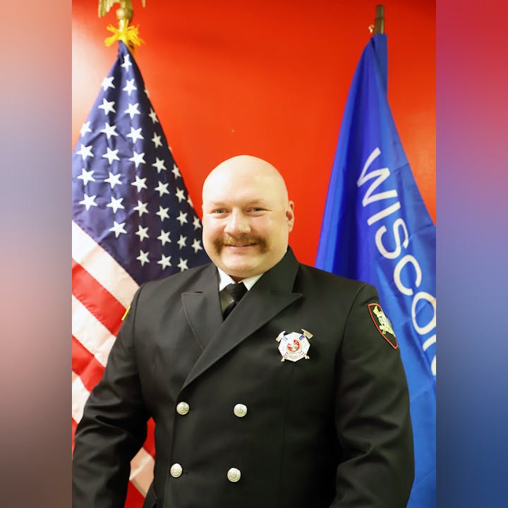 A smiling person in a formal uniform with badges in front of the US and Wisconsin flags.