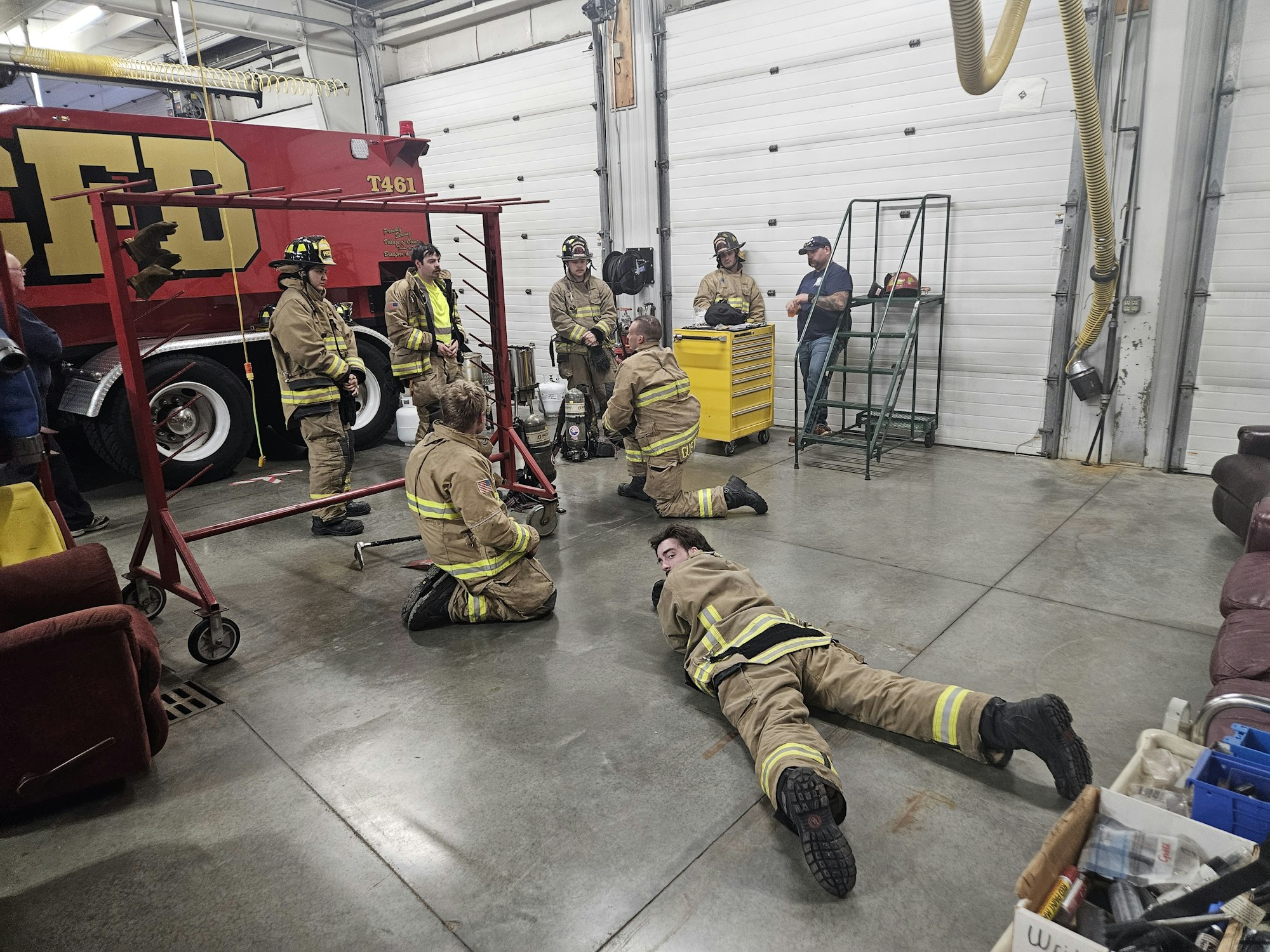 Firefighters in a garage with equipment and vehicles, some standing, others kneeling or lying on the floor.