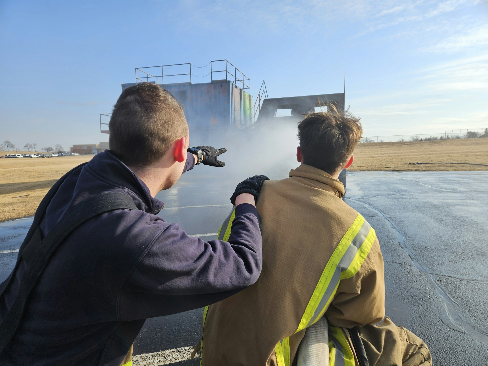 Two firefighters with a hose face a training structure emitting smoke outdoors.