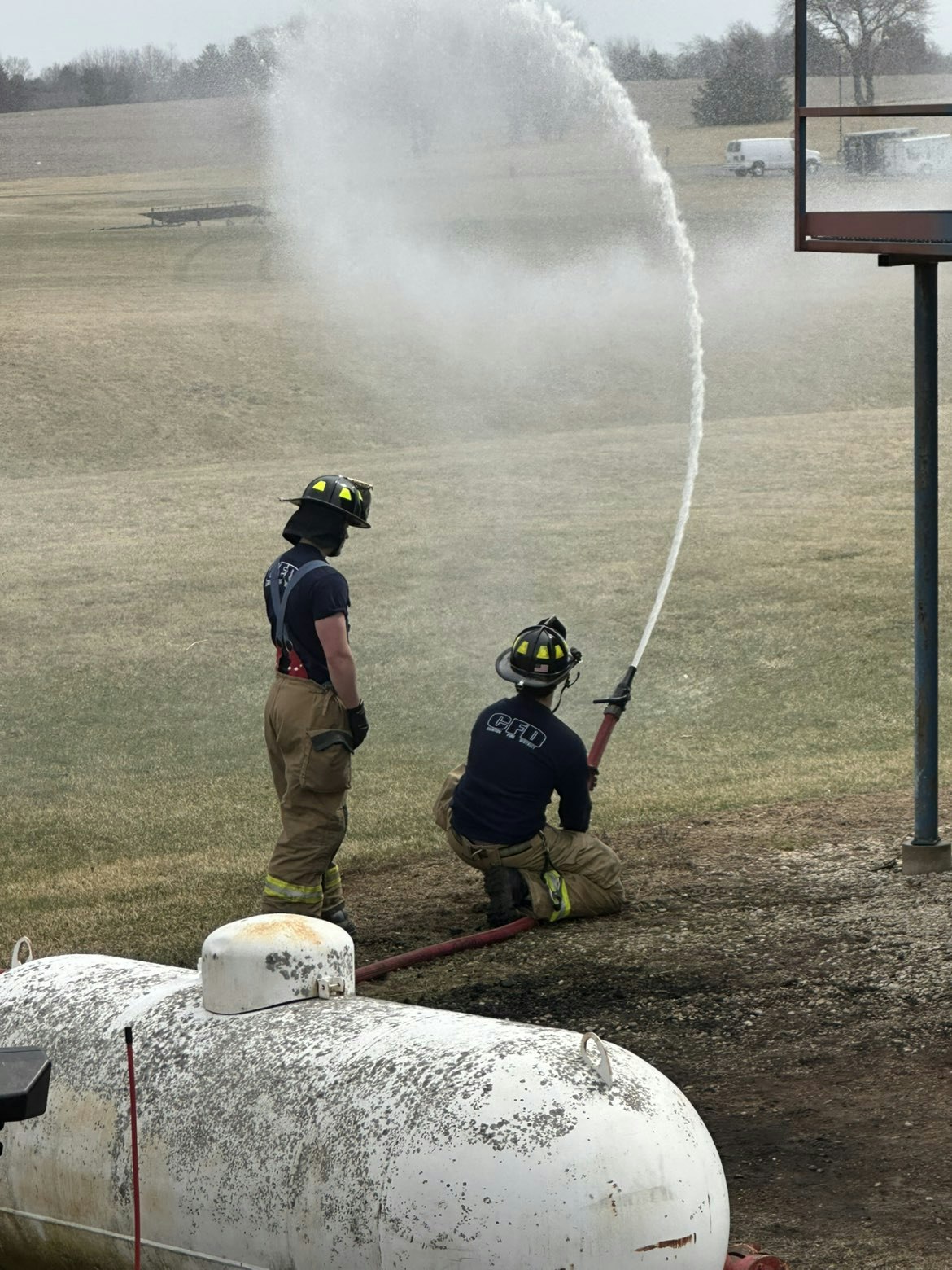 Two firefighters are using a hose to spray water, practicing techniques near a large tank in an open field.