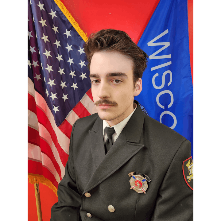 A person in uniform with badges stands in front of American and Wisconsin flags.