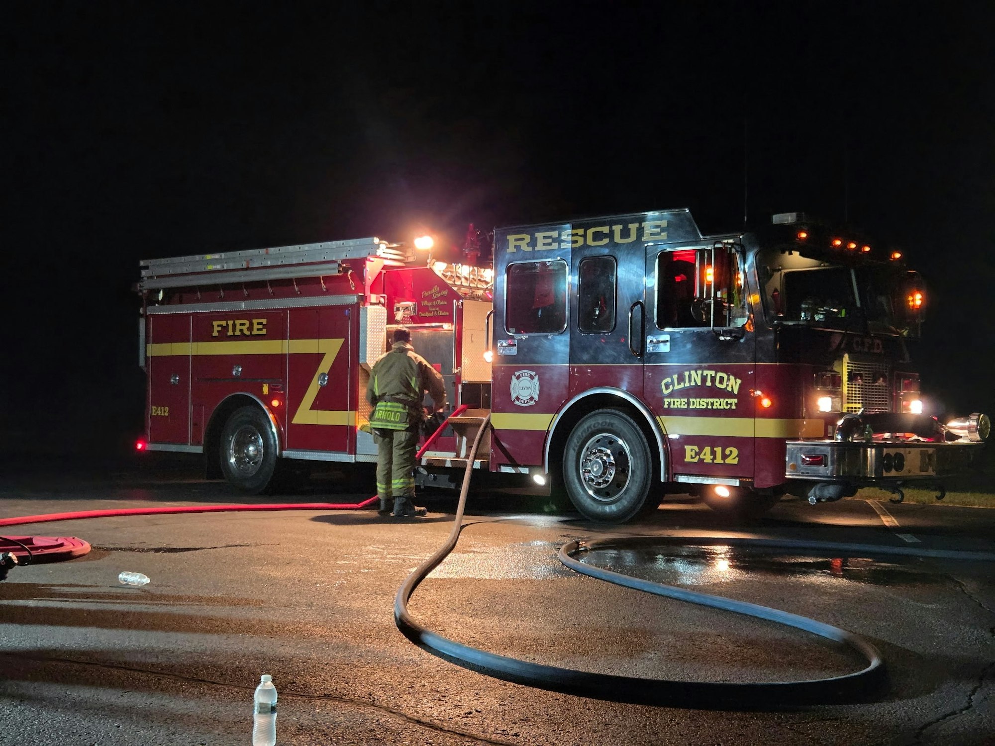 A fire truck labeled "Clinton Fire District" is parked at night, with a firefighter preparing equipment and hoses nearby.