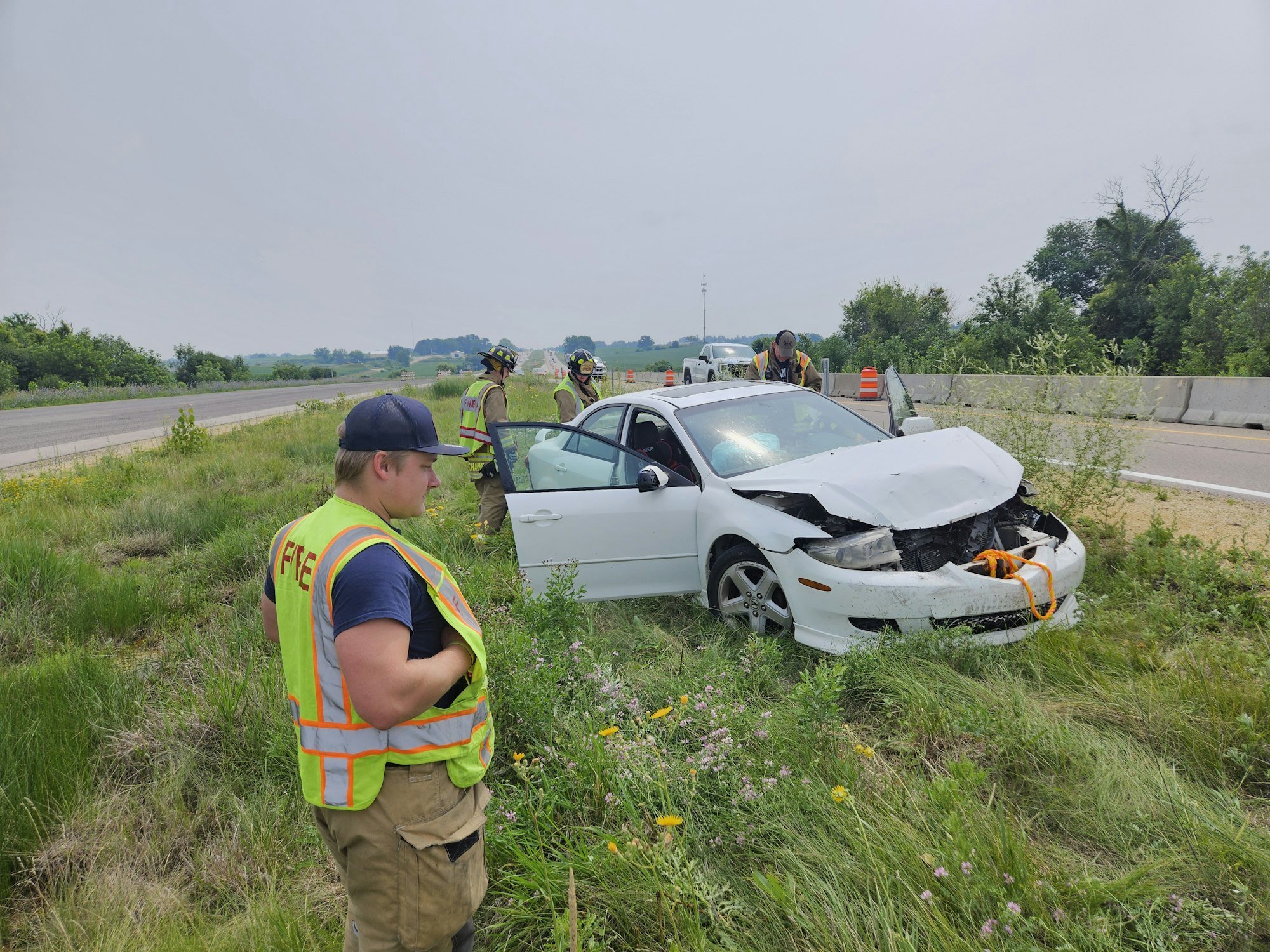 A damaged white car off the road, surrounded by emergency responders assessing the scene.