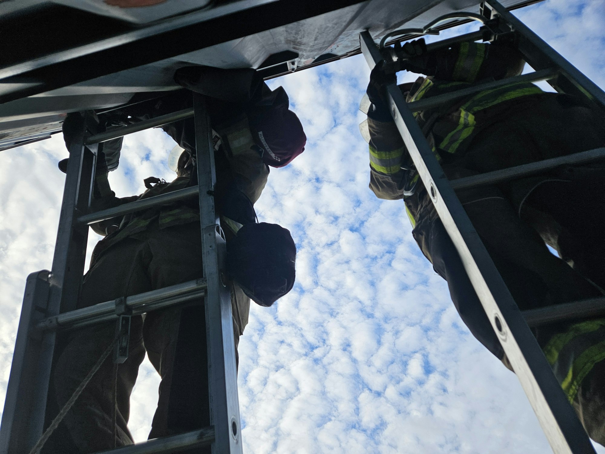 Two people in firefighting gear are on ladders under a cloudy sky.