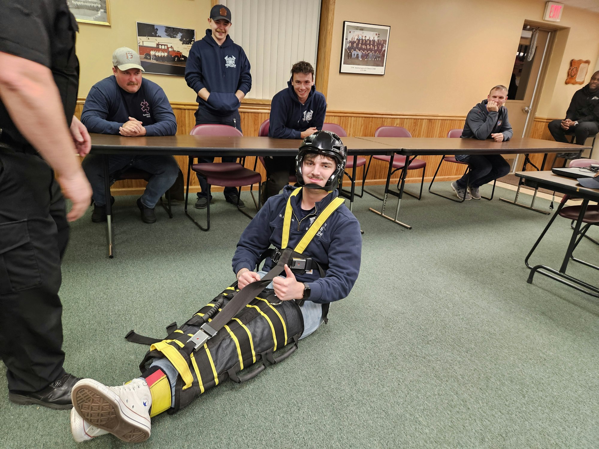A person in safety gear is sitting on the floor, while others observe from a table in a training or instructional setting.