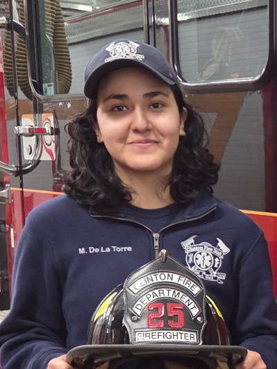 A young woman stands in front of a Clinton Fire District truck, holding a firefighter helmet, showcasing pride in her role.