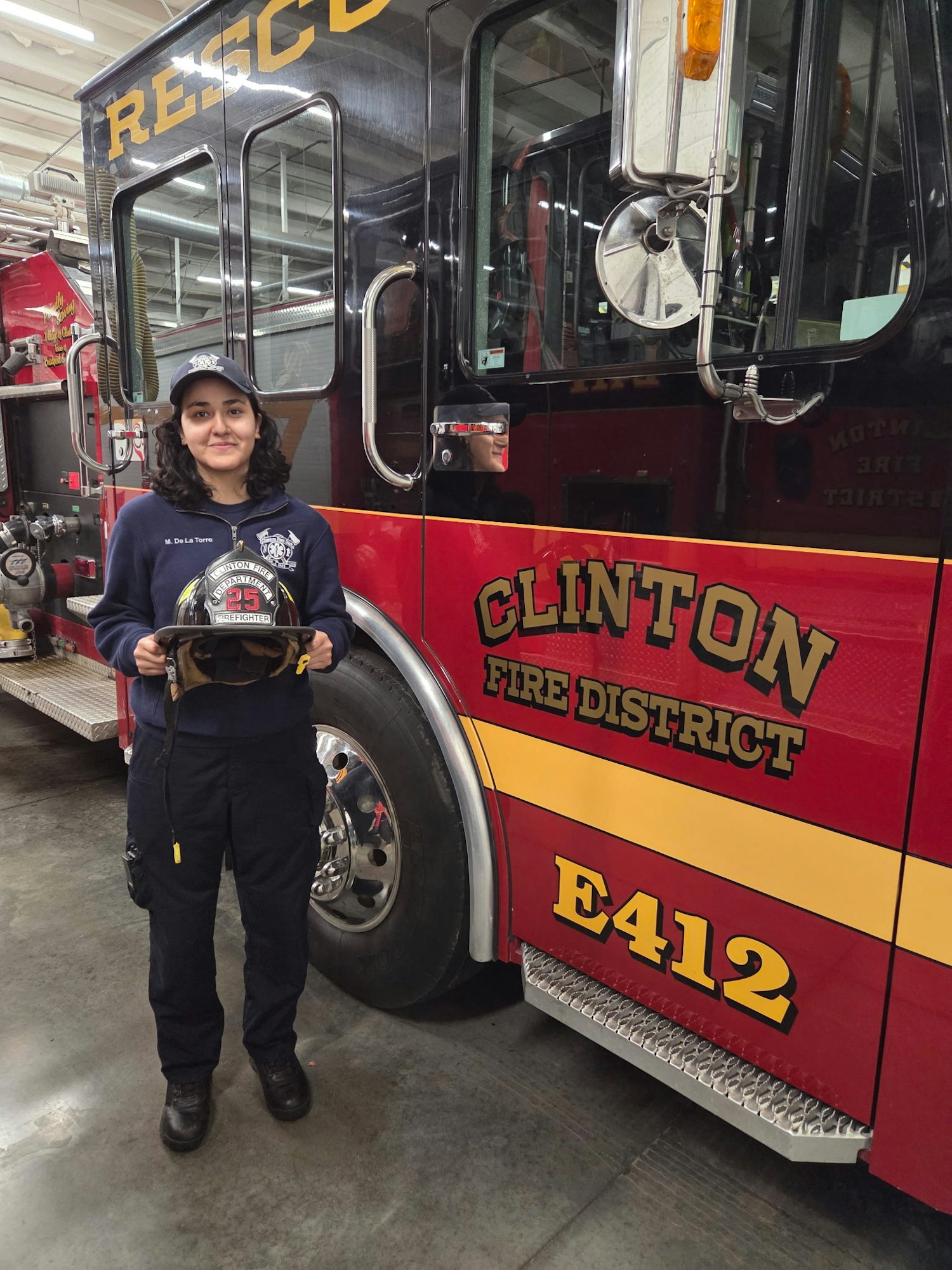 A young woman stands in front of a Clinton Fire District truck, holding a firefighter helmet, showcasing pride in her role.