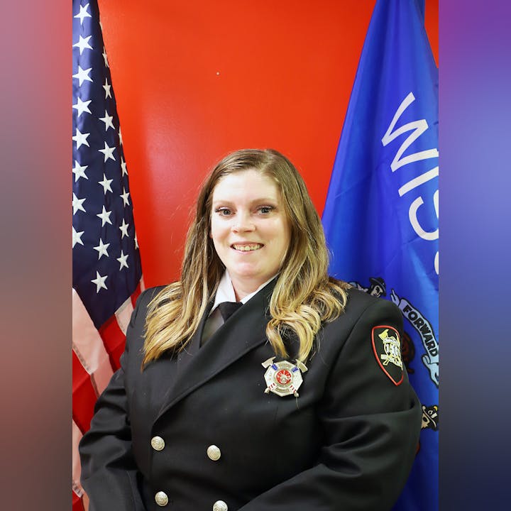 A woman in a formal uniform with badges in front of the US flag and another flag, on a red background.
