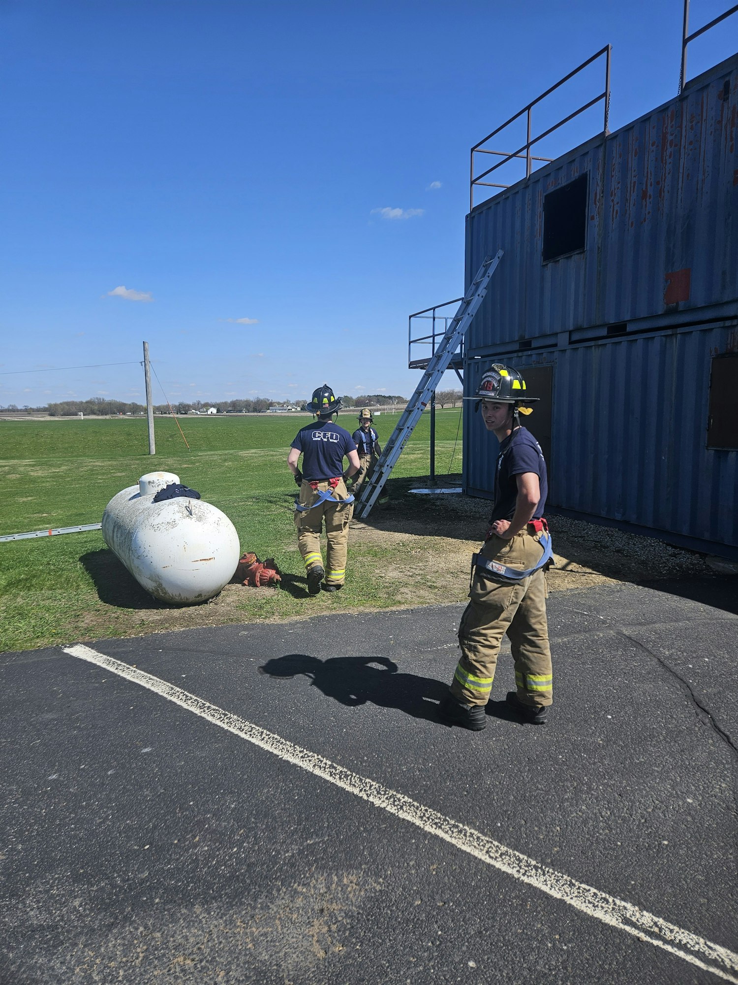 Two firefighters stand by a ladder and a training structure with clear skies.