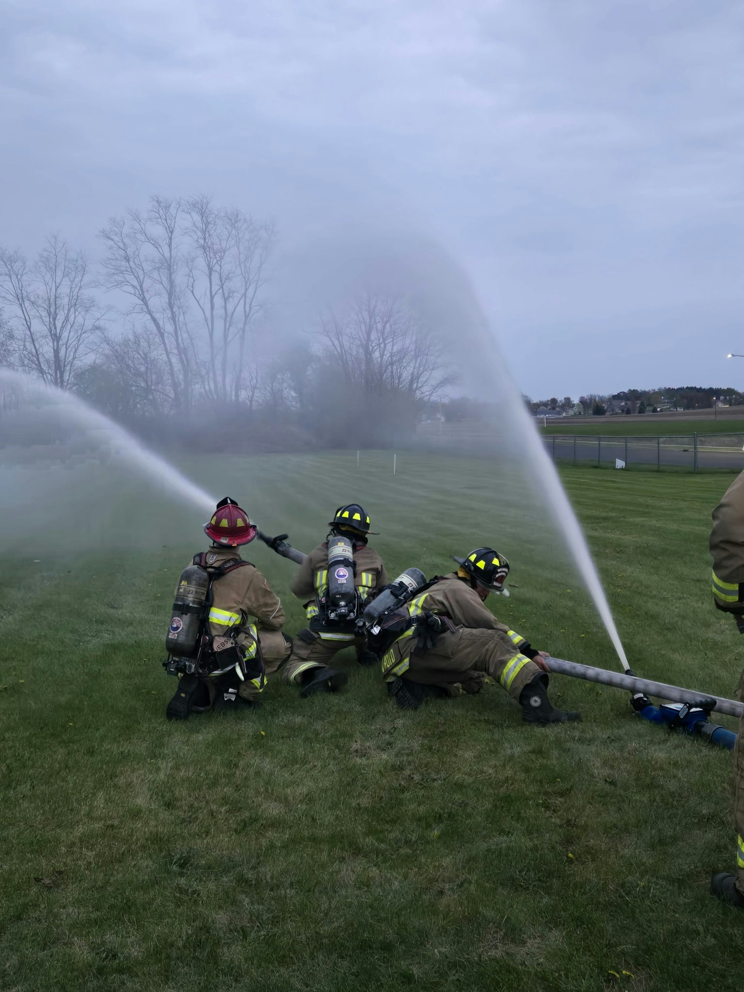 Firefighters in gear spray water hoses on a grassy field, simulating a firefighting exercise.