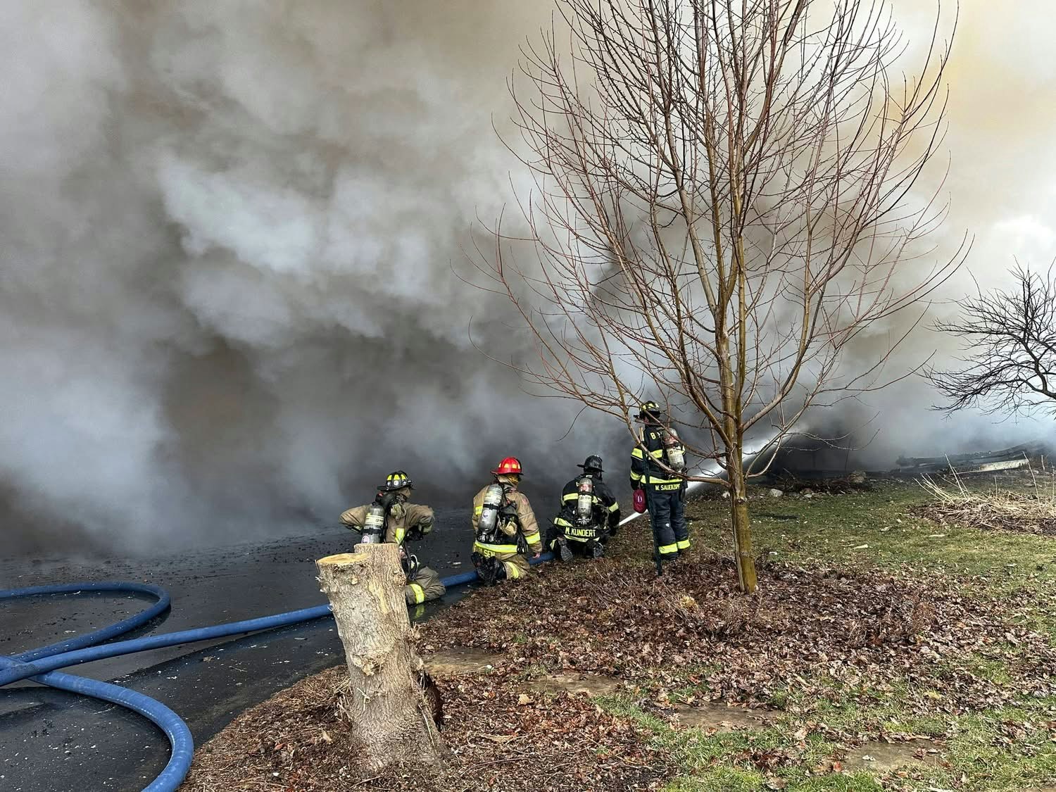 Firefighters respond to a blaze, surrounded by thick smoke and hoses, working to control the situation near a tree.