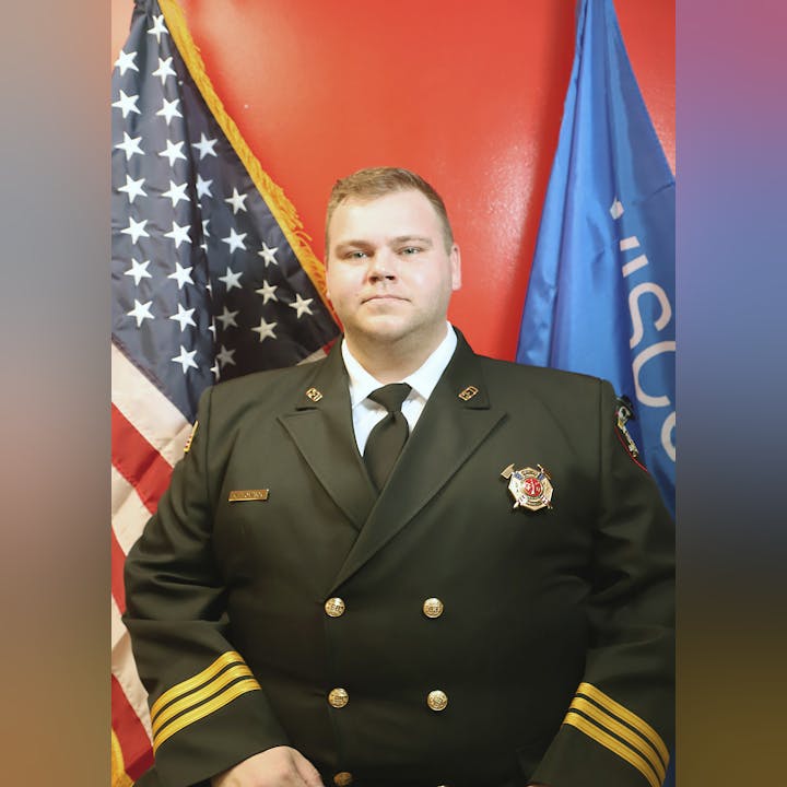A person in a fire department uniform with badges in front of the US and fire department flags.