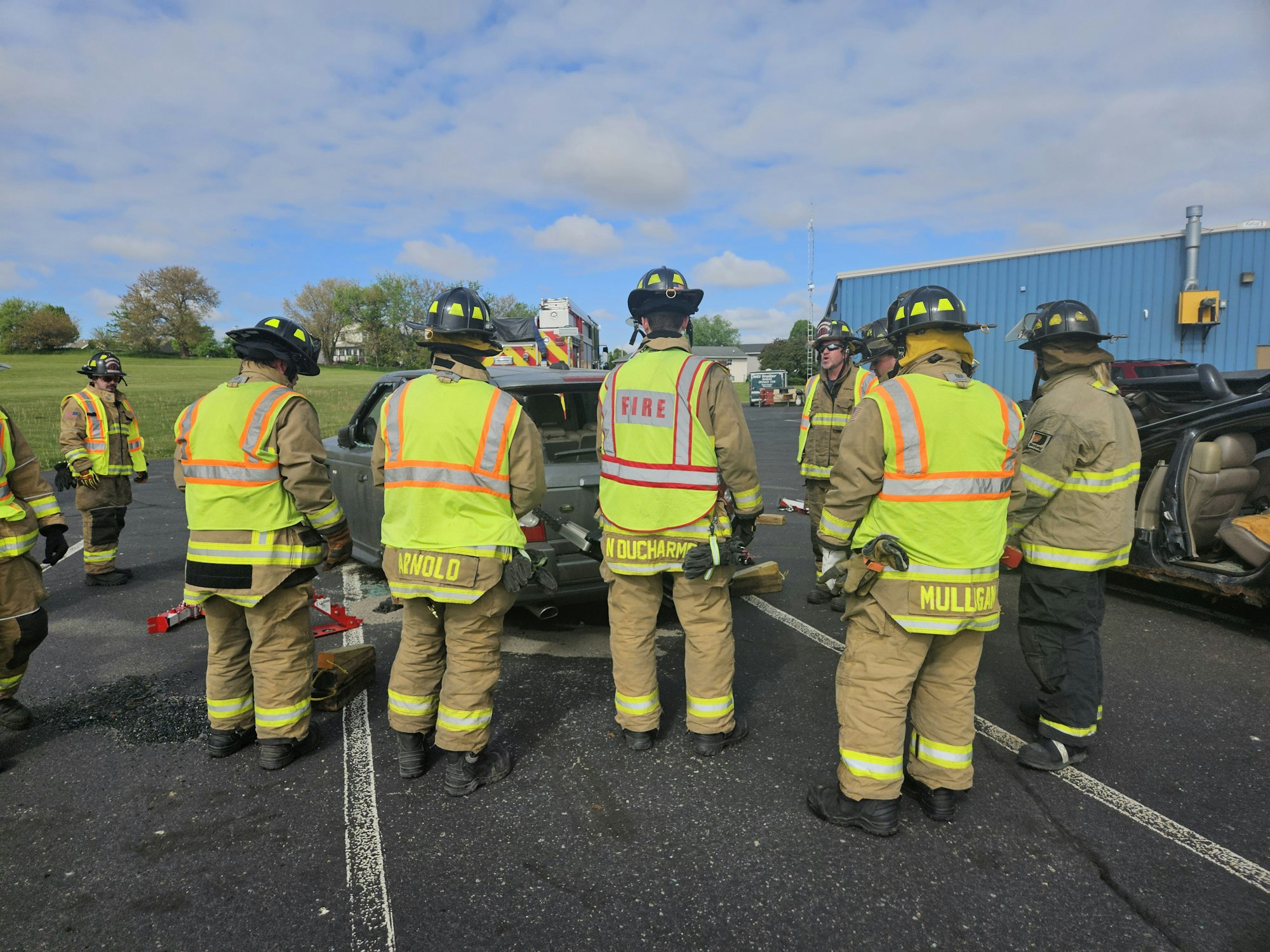 Firefighters in gear and yellow vests gather near a car, possibly for training or an emergency response.