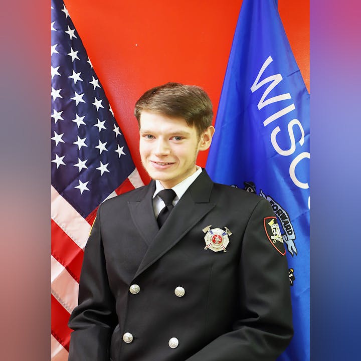 A man in a dress uniform with badges in front of the US flag and another blue flag.