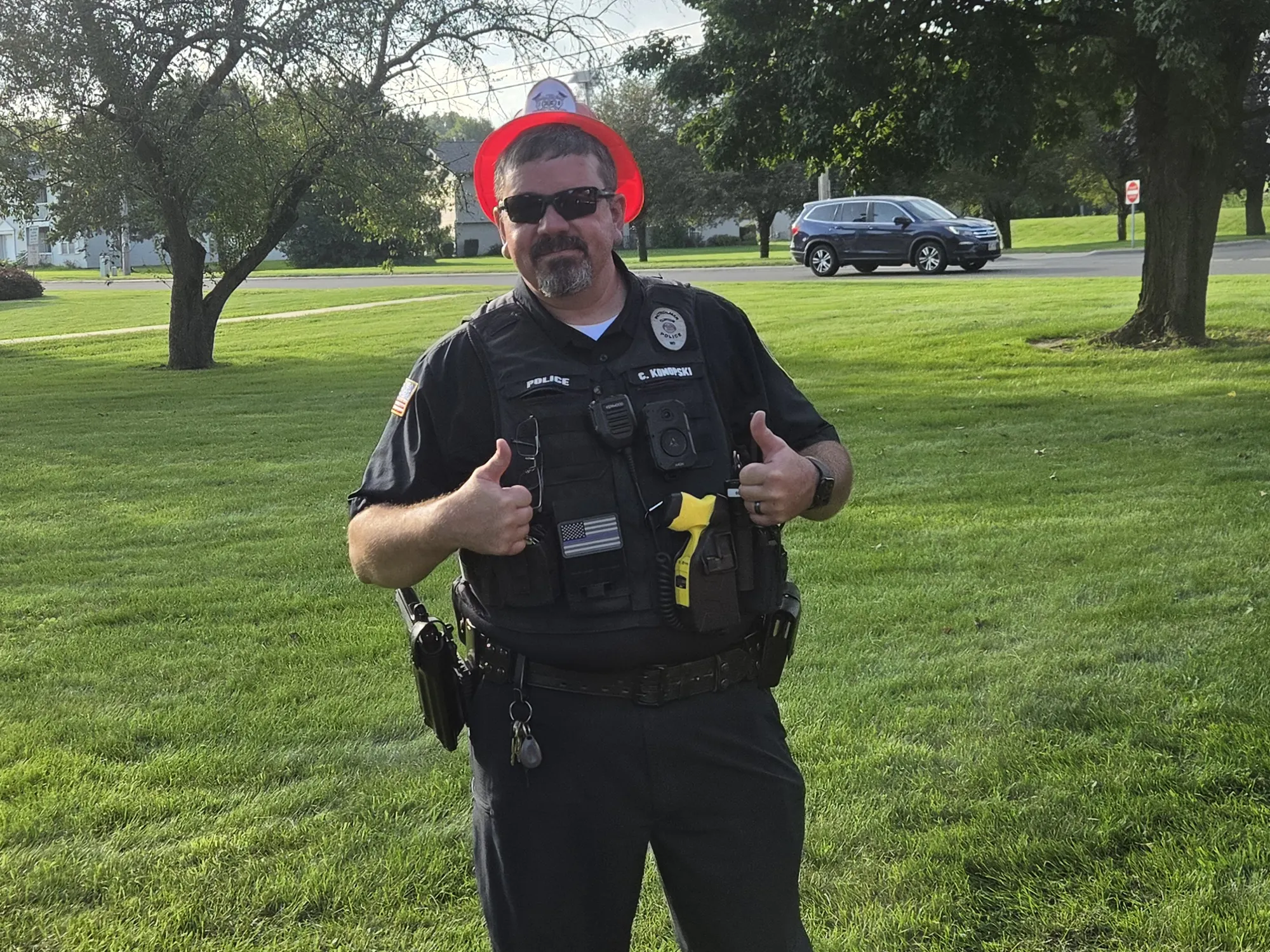 An officer stands on grass, giving a thumbs-up, wearing sunglasses and a badge, with trees and a street in the background.