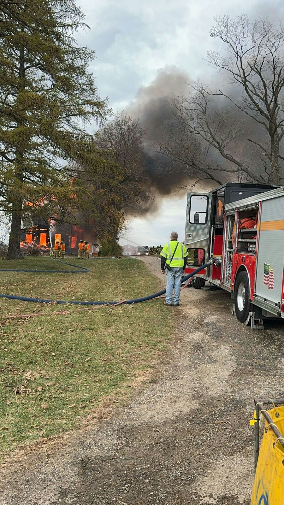 A firefighter observes as a house fire rages, with a fire truck and hose in the forefront.