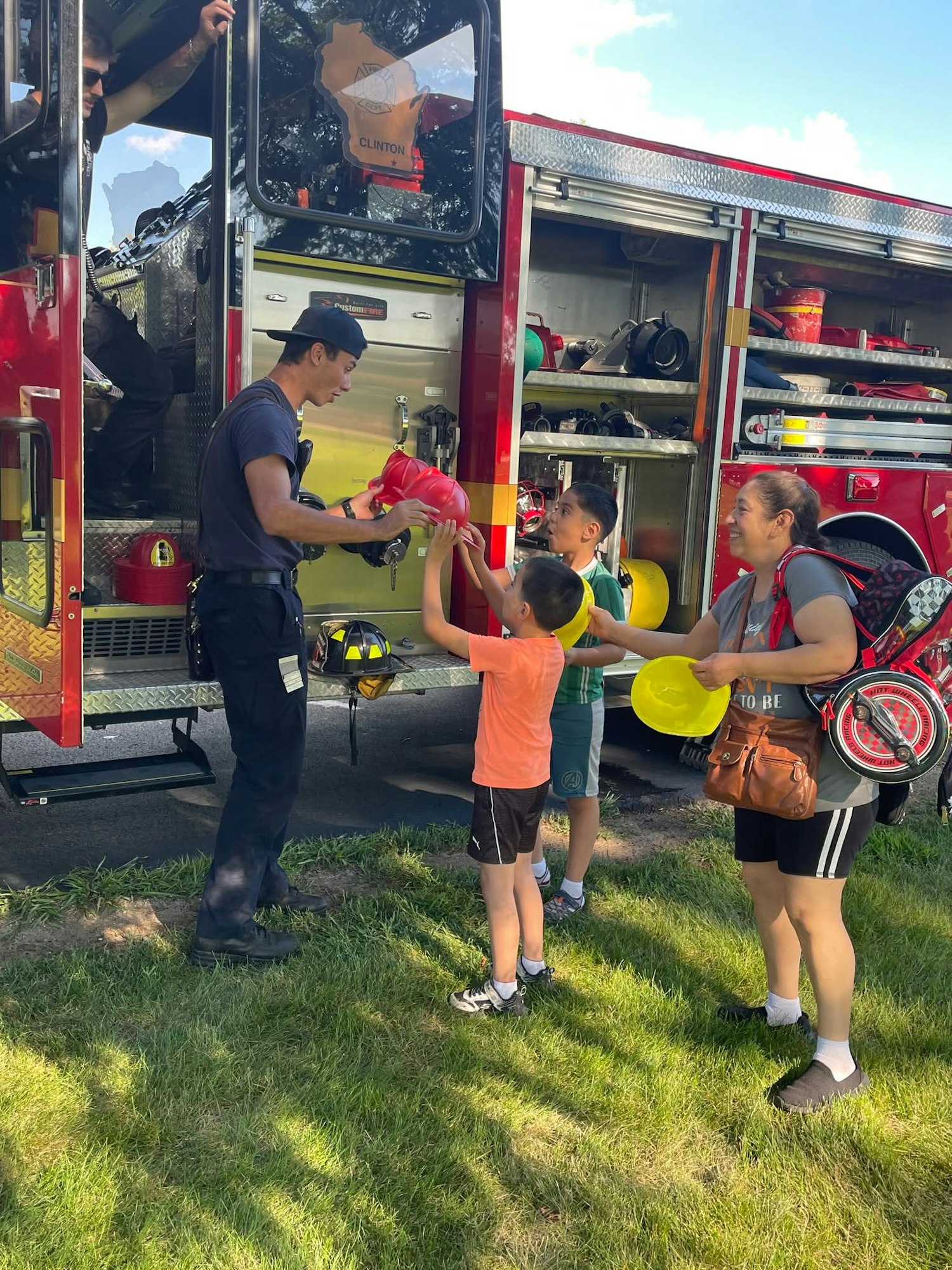 A firefighter hands a red helmet to a child near a fire truck, with more children and a woman nearby.
