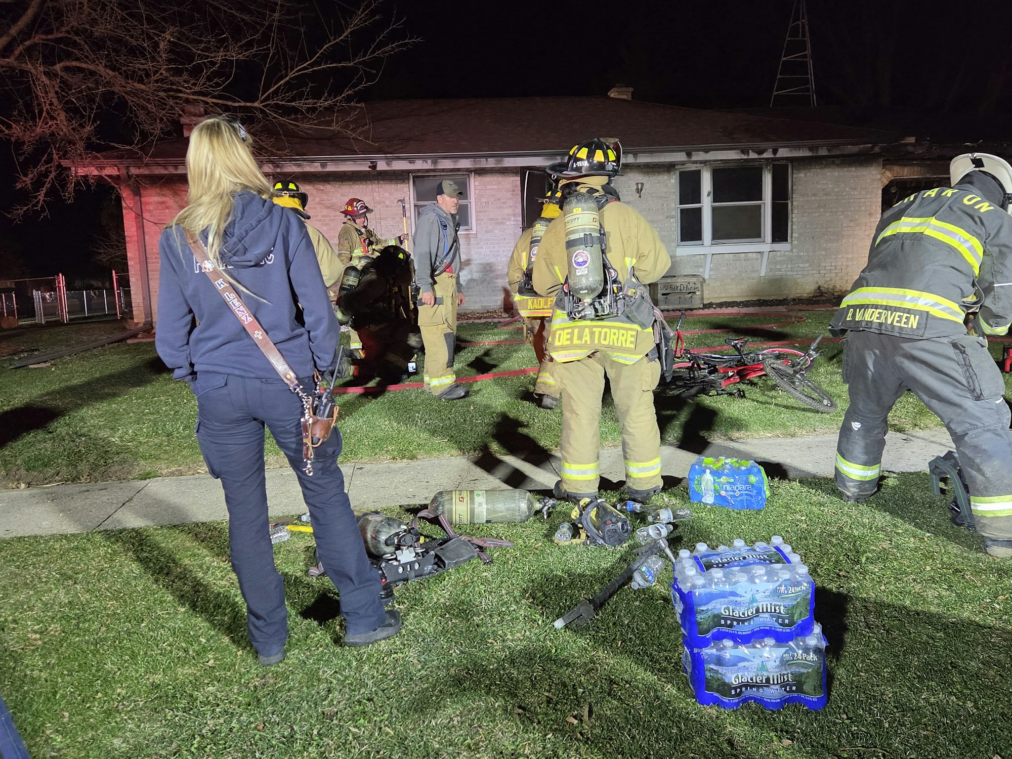 Firefighters respond to an incident at a home, with equipment and bystanders present. It's nighttime, and emergency lights are on.