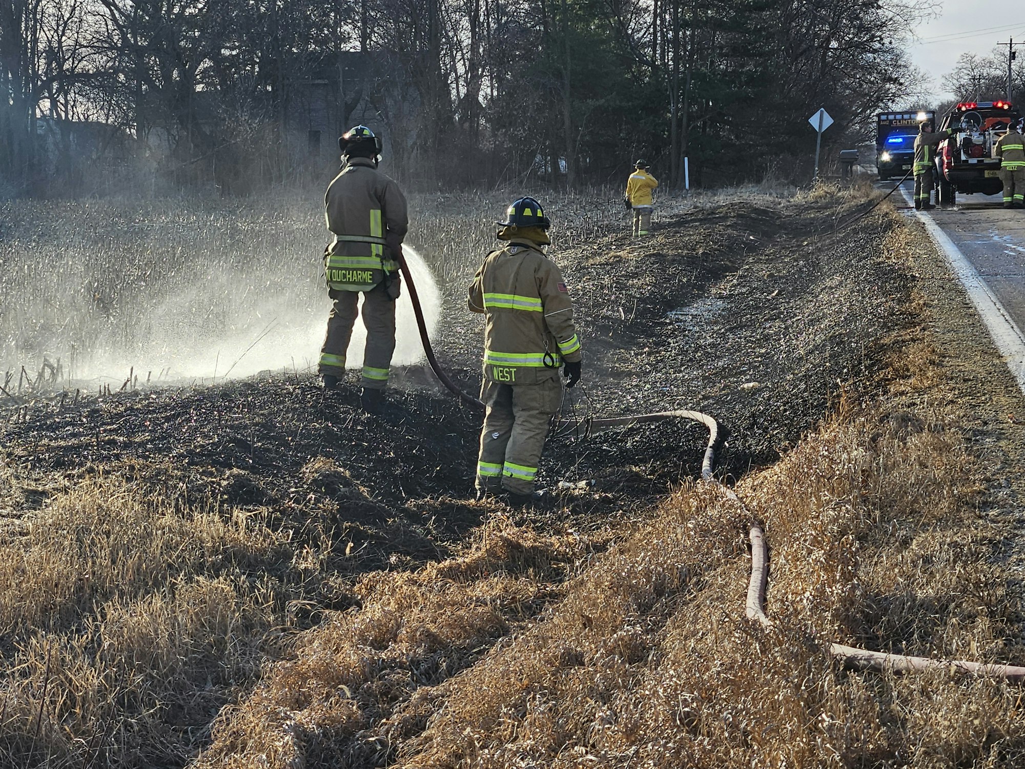 Firefighters extinguish a roadside grass fire using hoses, while emergency vehicles are parked nearby.