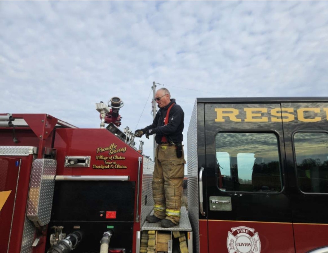 A firefighter operates equipment on a fire truck labeled "RESCUE" under a cloudy sky.