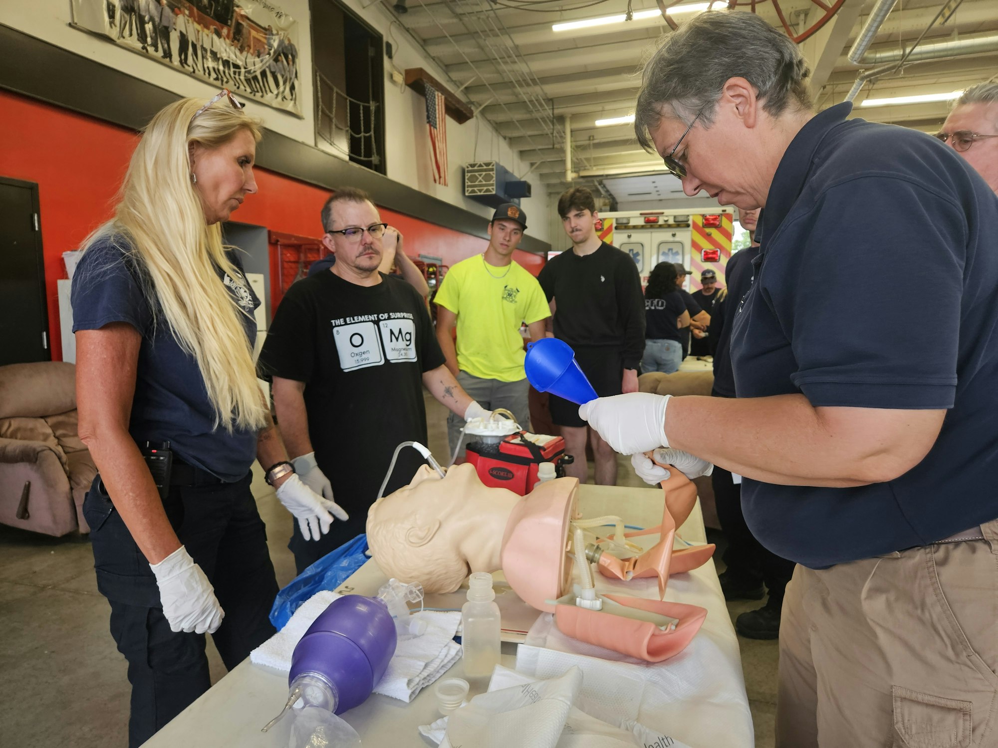 People in a training session with medical equipment and a mannequin on a table.