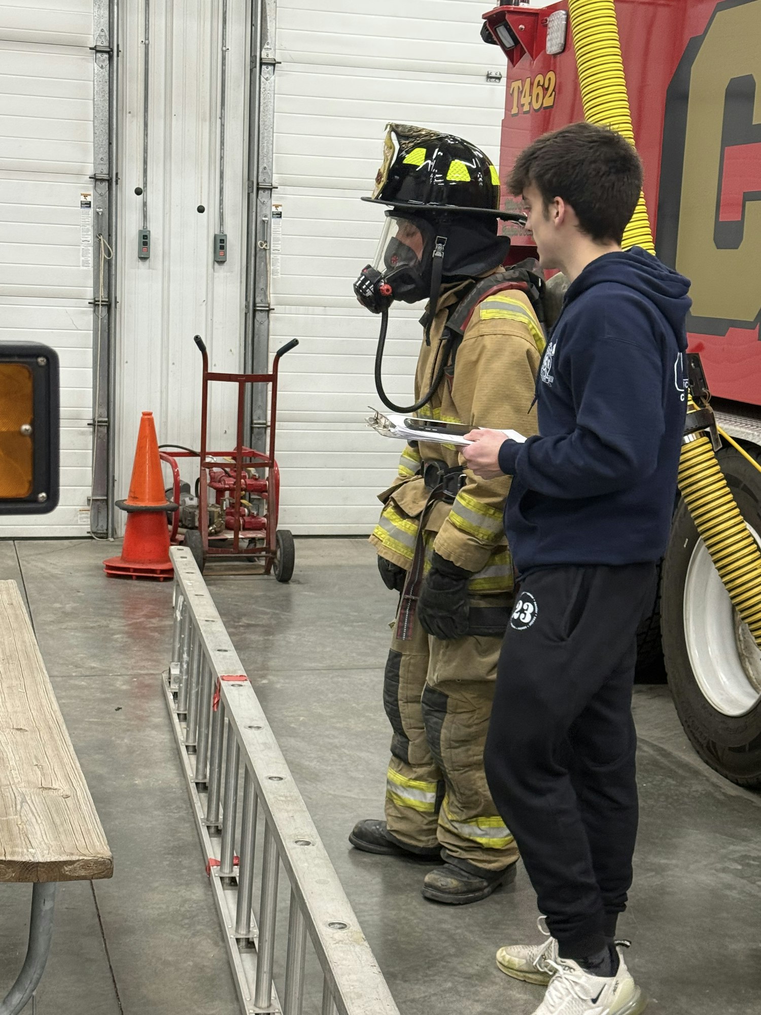 A person in a firefighter suit stands next to another individual holding a tool, inside a fire station with equipment visible.