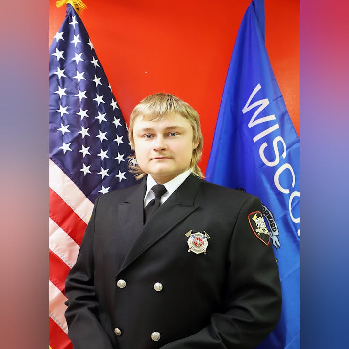 A person in a formal uniform with badges, between the US and Wisconsin flags.