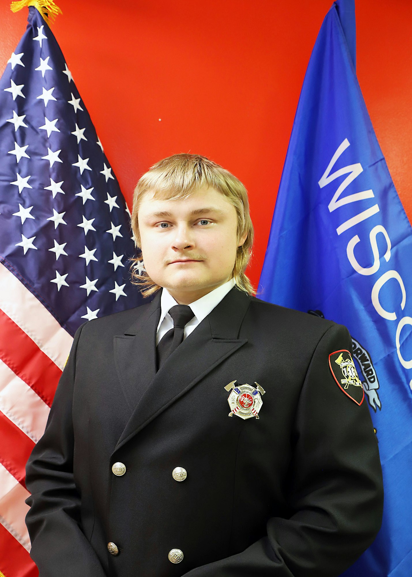 A person in a formal uniform with badges, between the US and Wisconsin flags.