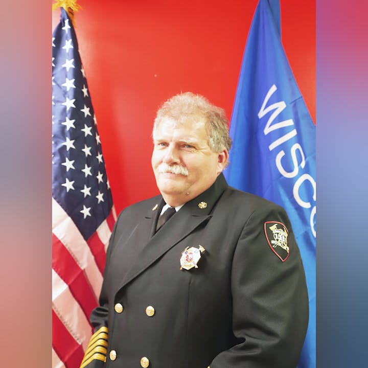 A person in firefighter uniform with badges, standing in front of the US and Wisconsin flags.