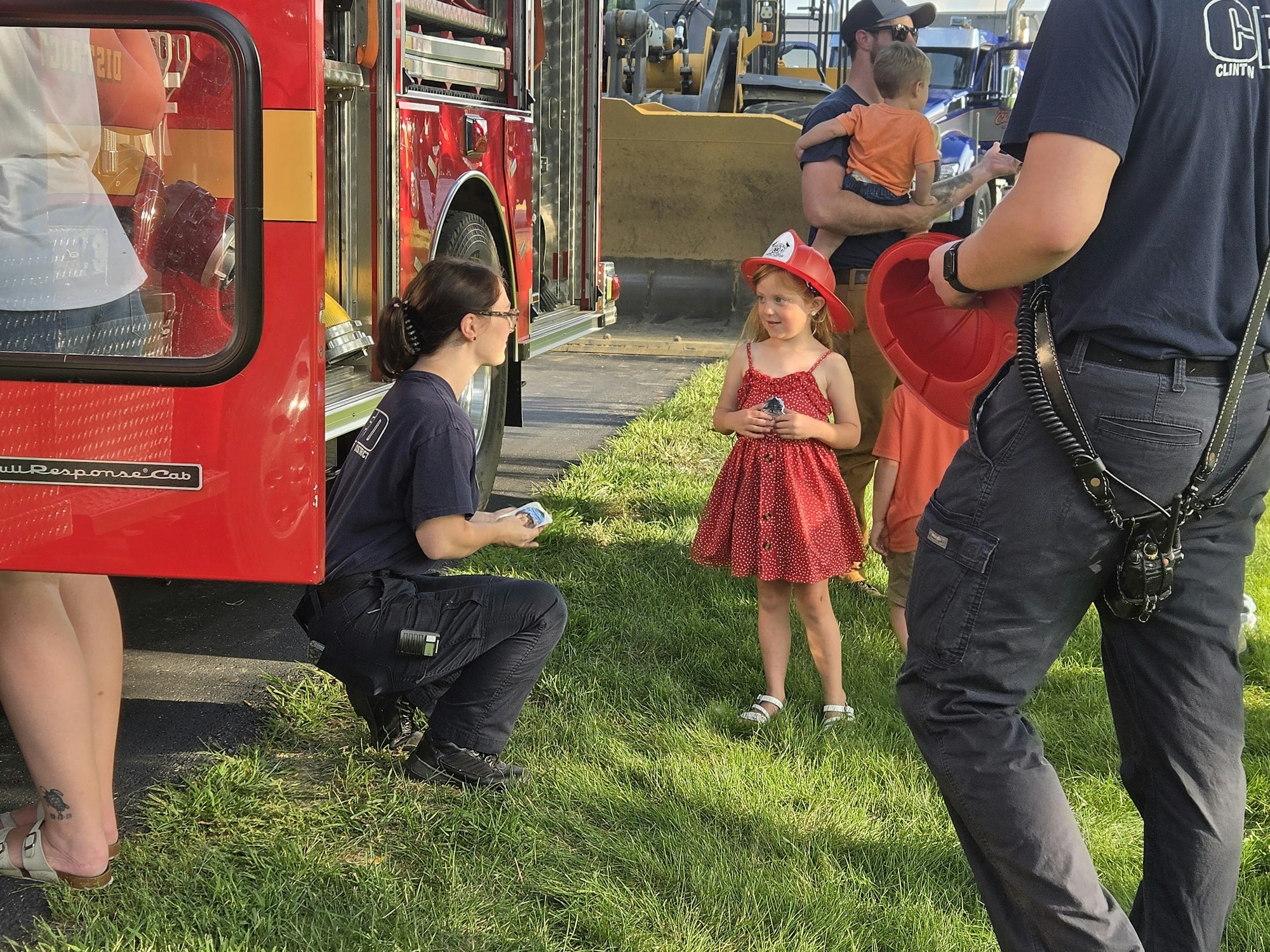A young girl in a red dress and firefighter hat interacts with a woman near a fire truck, surrounded by other people.