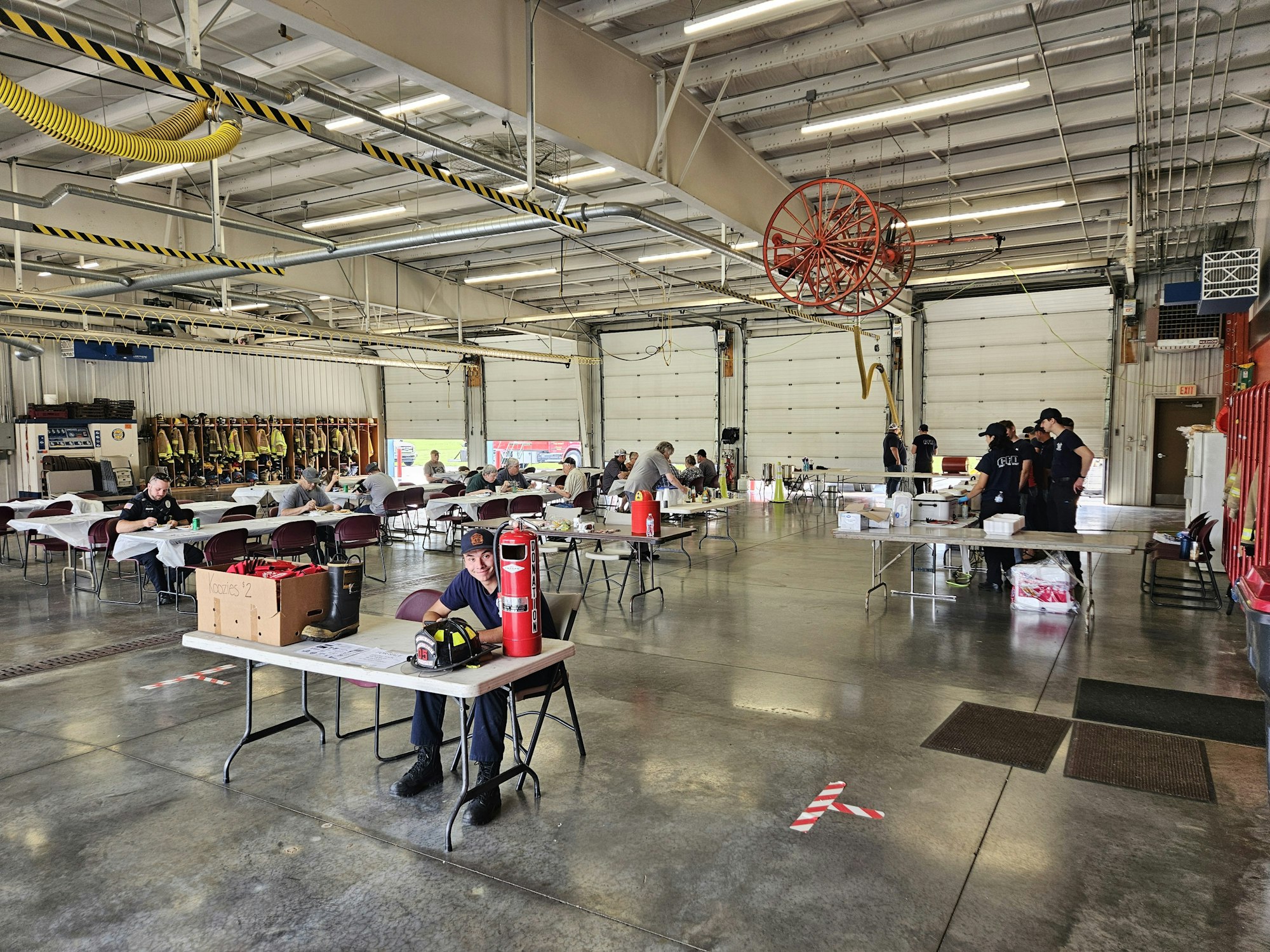 A large indoor space with tables and chairs, people sitting and standing, fire station gear in background.