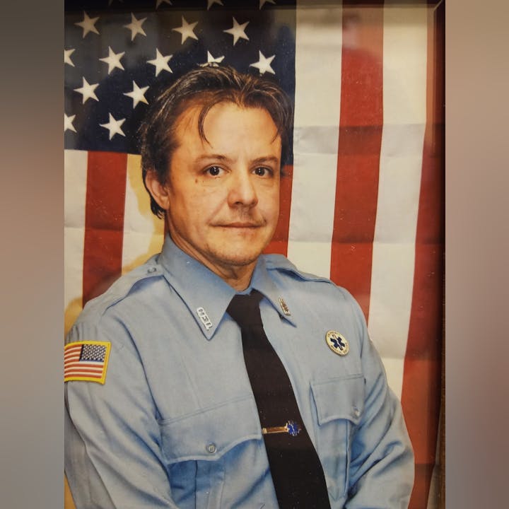 A person in a blue uniform with patches and badges, standing before an American flag background.