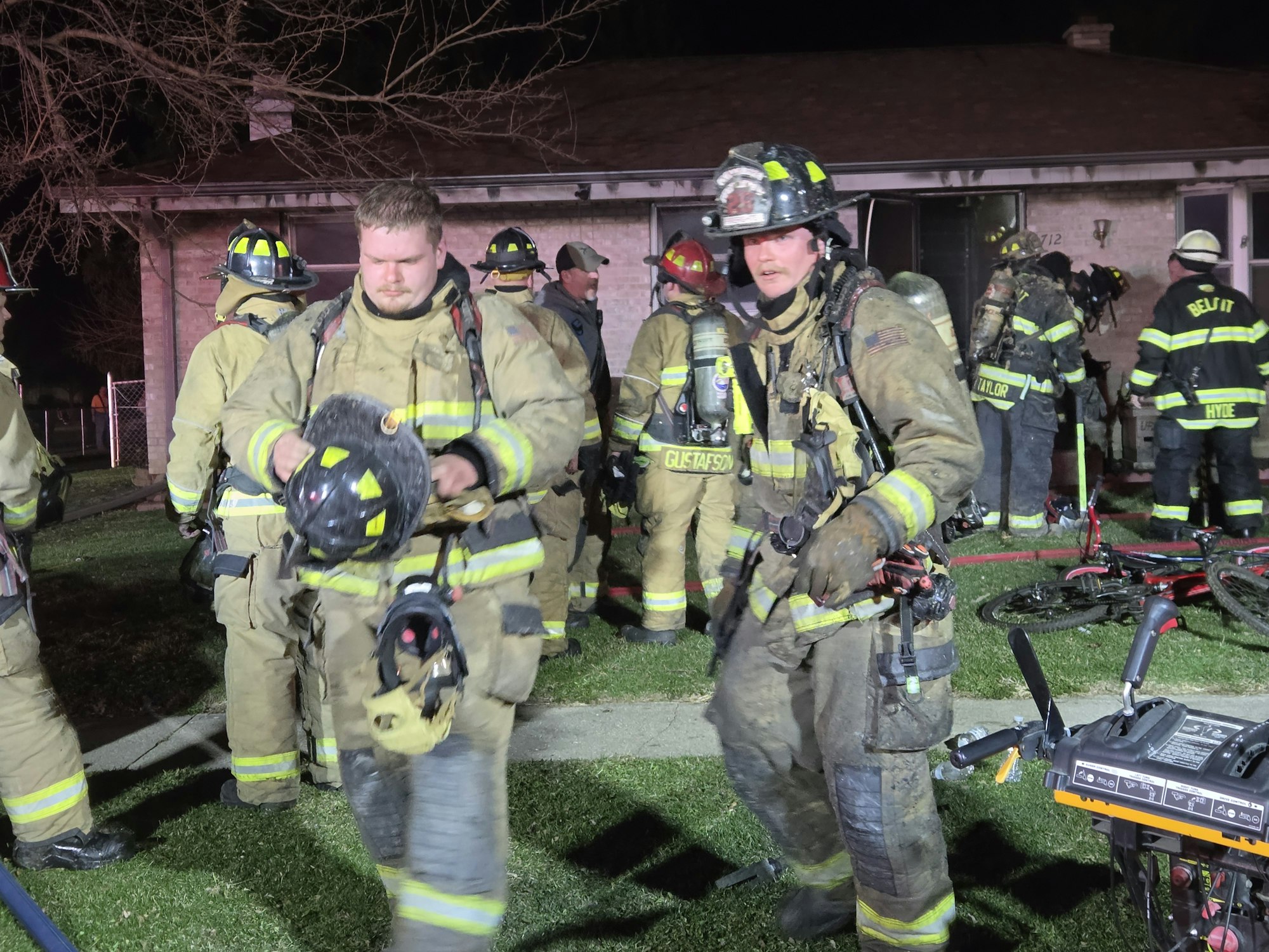 Firefighters in gear responding to a blaze outside a house, with equipment and bicycles visible nearby.