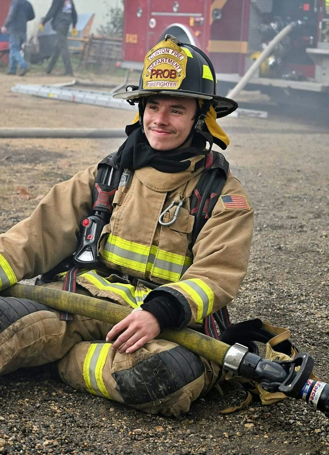A smiling firefighter sitting on the ground in full gear, holding a hose, with a fire truck in the background.