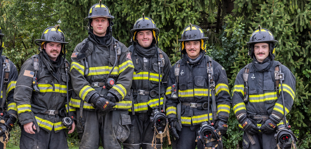 A group of five firefighters in gear posing for a photo outdoors, showcasing camaraderie and readiness for duty.