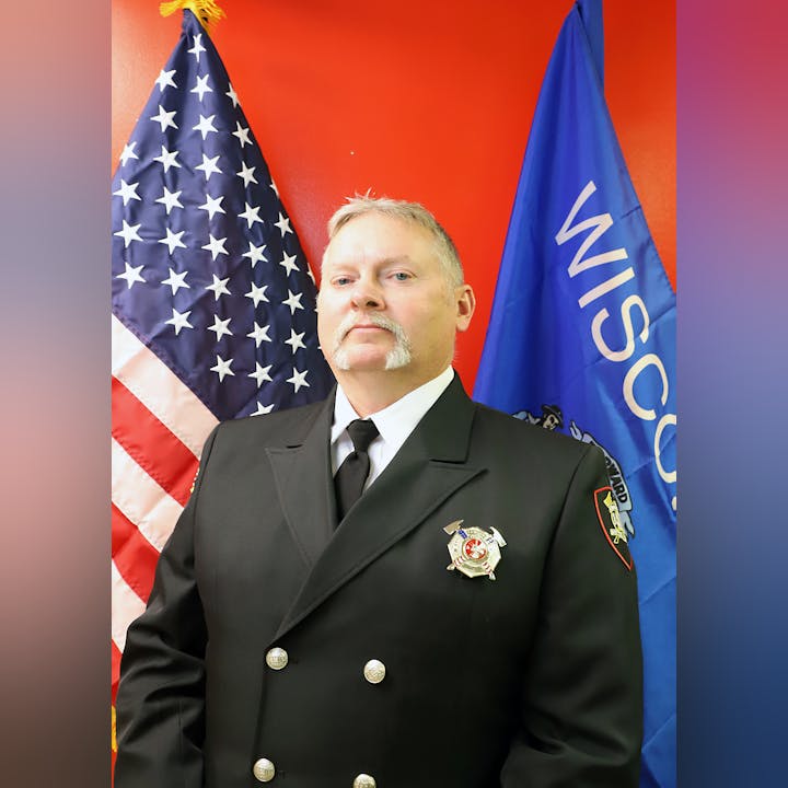 A man in a uniform with badges in front of the US and Wisconsin flags.