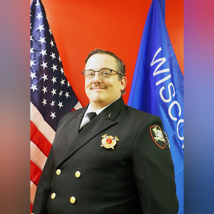 A person in a formal uniform with badges stands between the US and Wisconsin flags.