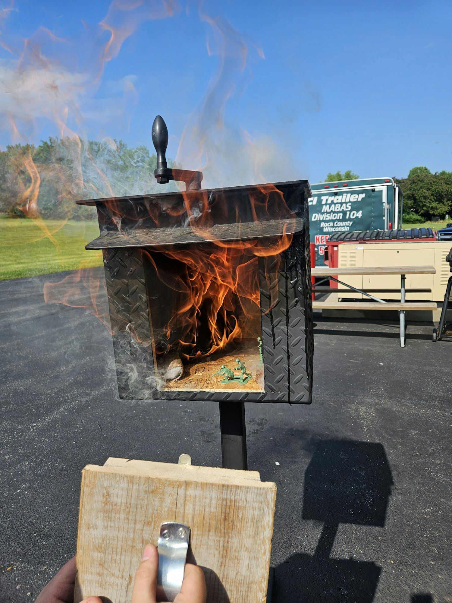 A mailbox on fire with smoke, person's hand holding a wooden board, and a parked truck in the background.