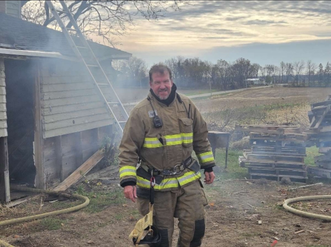 A firefighter in full gear standing before a building with a ladder and smoke in the background.