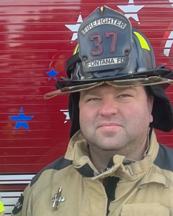 Two firefighters in uniform pose in front of a fire truck, showcasing their gear and a decorative background.