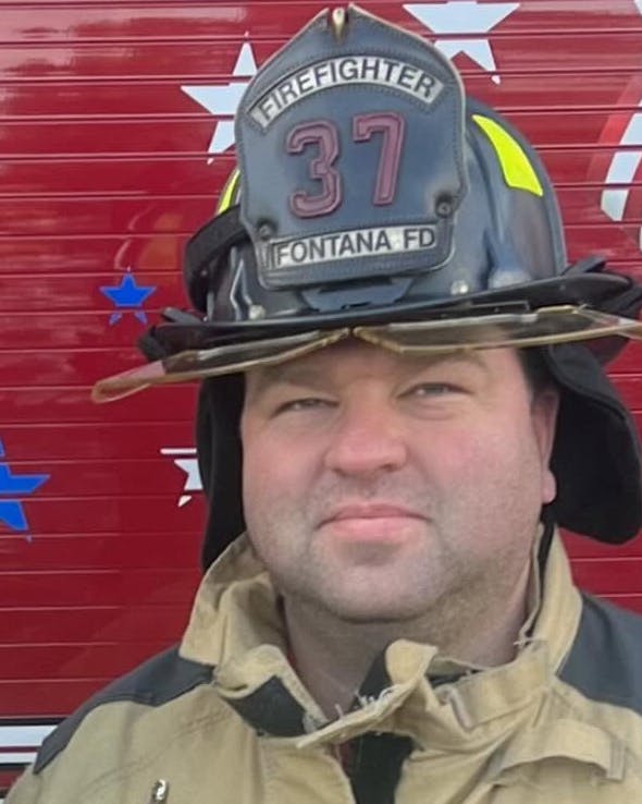 Two firefighters in uniform pose in front of a fire truck, showcasing their gear and a decorative background.