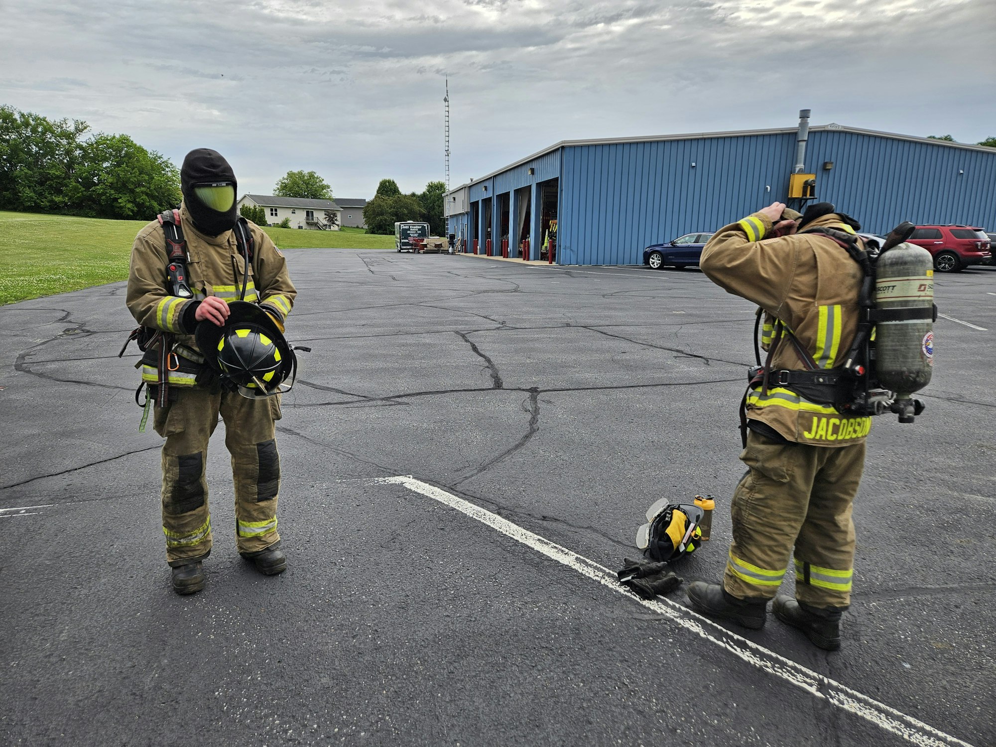 Two firefighters in gear outside a fire station. One holds a helmet; the other adjusts equipment.