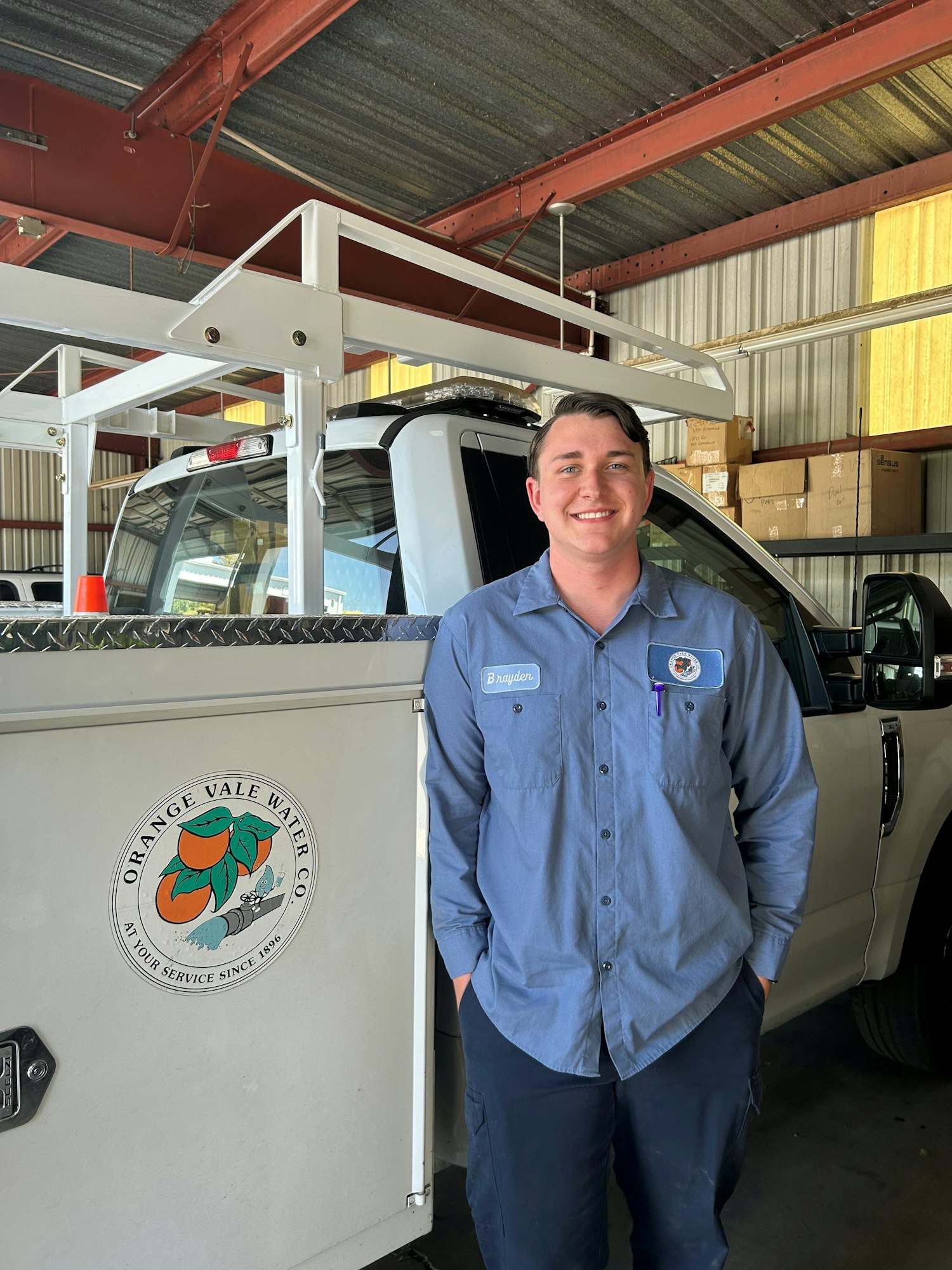 Person in a blue shirt smiling next to a truck with an "Orange Vale Water Co." logo in a garage.